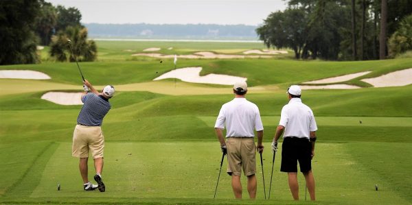 Golfer in striped shirt swings a drive as two men watch on a lush golf course with sand traps in the background.